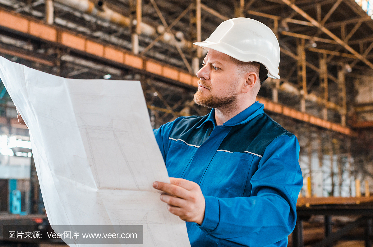 一個(gè)穿著工作服、戴著白色頭盔的工人正在檢查一個(gè)金屬工廠背景上的圖紙。建筑工人身體很好。男工程師